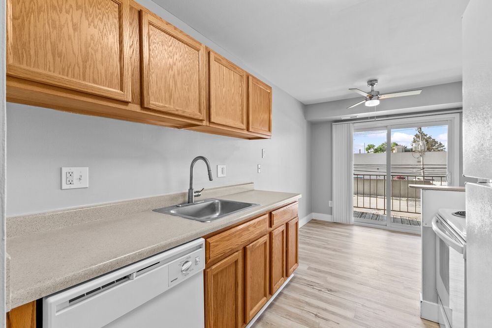 Modern kitchen featuring oak cabinets, white dishwasher, single basin sink, and sliding glass door to balcony.