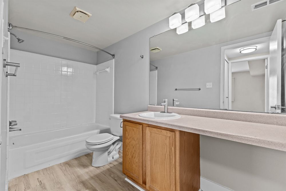 Clean bathroom with oak vanity, large mirror, white tub-shower combo, toilet, and wood-look flooring.