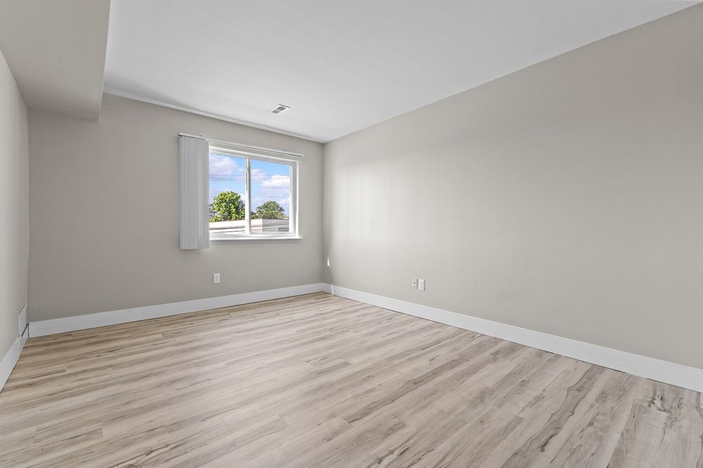Bright empty bedroom with light wood laminate floors, large window, and neutral gray walls.