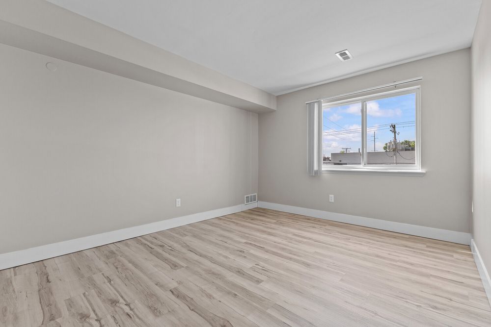 Empty room with light hardwood floors, gray walls, and a single window with natural light in a modern apartment.