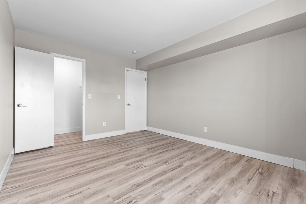 Bright empty apartment bedroom with light gray walls, two white doors, and light wood-look flooring.