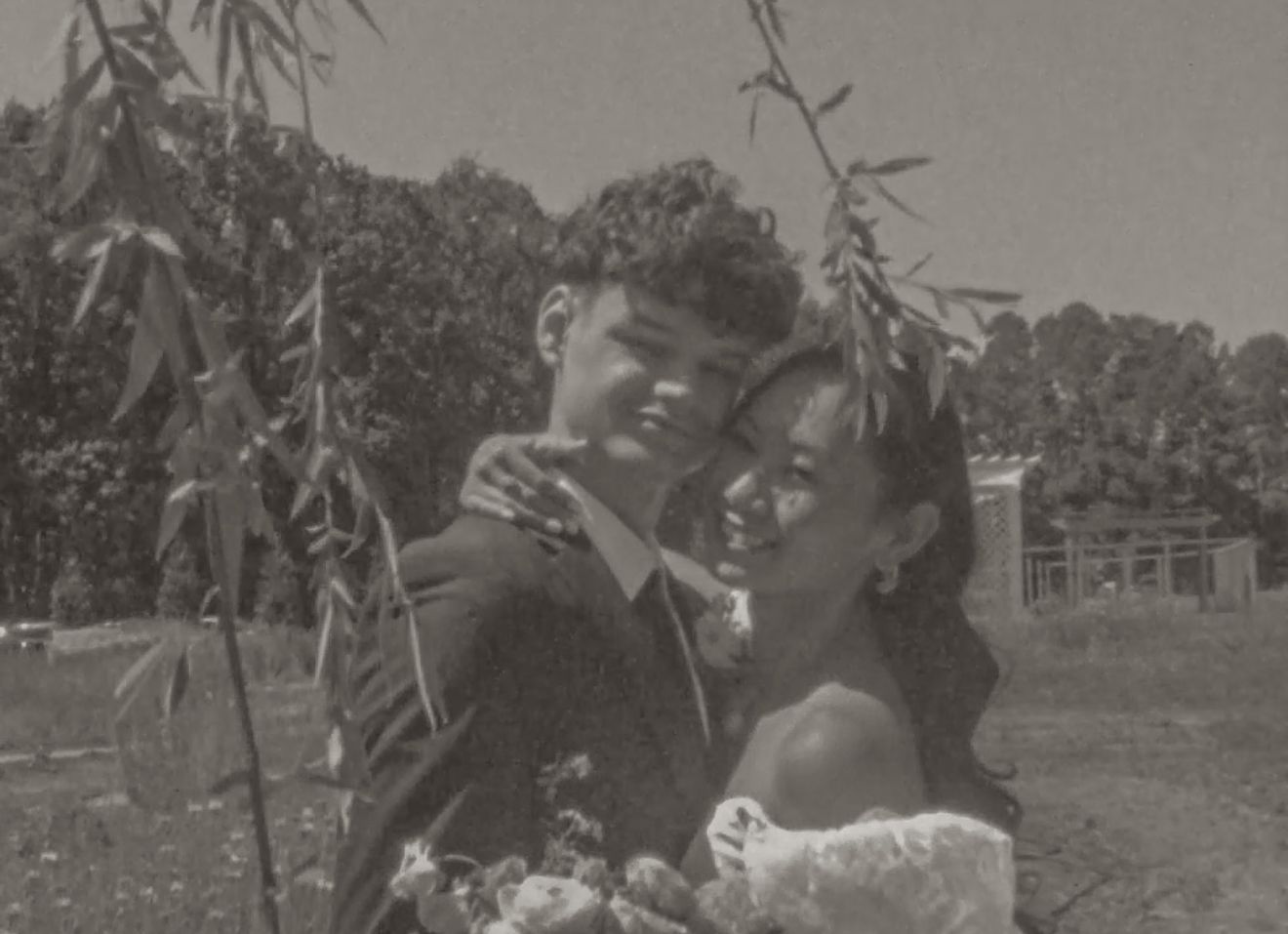 Black and white vintage wedding photo of a couple embracing outdoors, bride in off-shoulder gown, groom in suit.