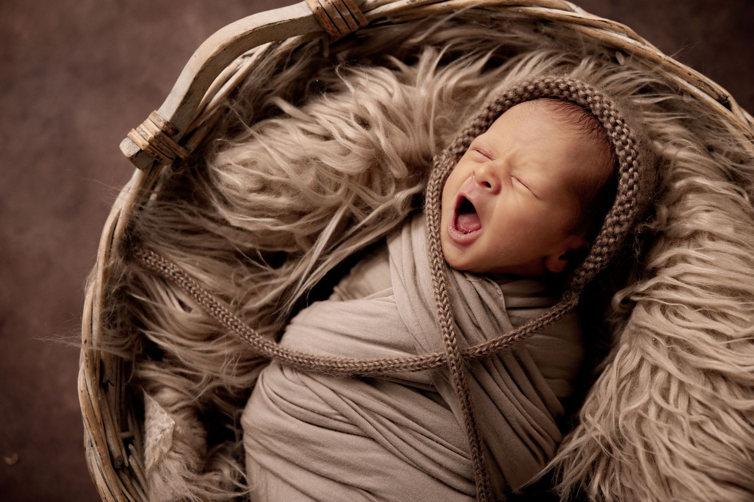 Newborn baby yawning, swaddled in beige wrap, wearing a knit bonnet, resting in a rustic basket with fur lining.