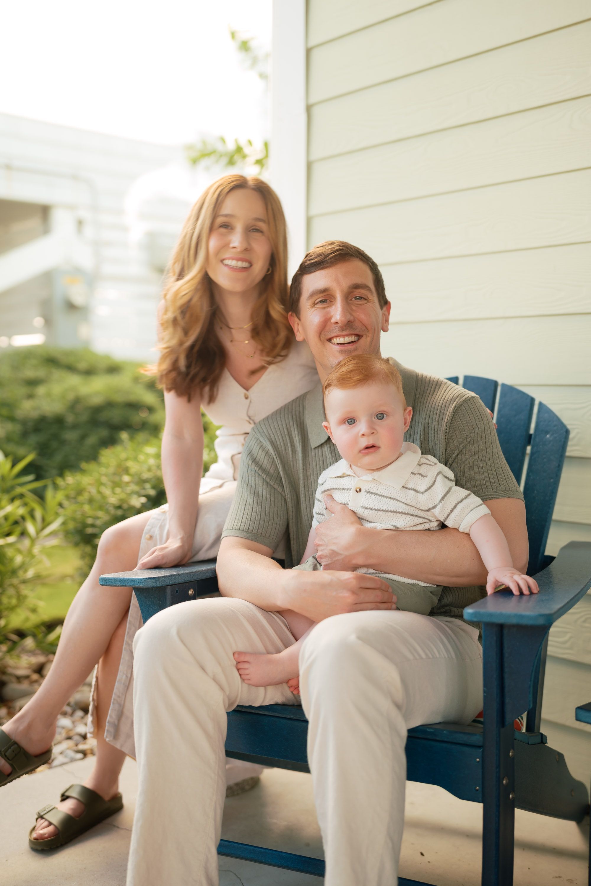 Happy young family with baby sitting on blue Adirondack chair on home porch, smiling couple enjoying parenthood.
