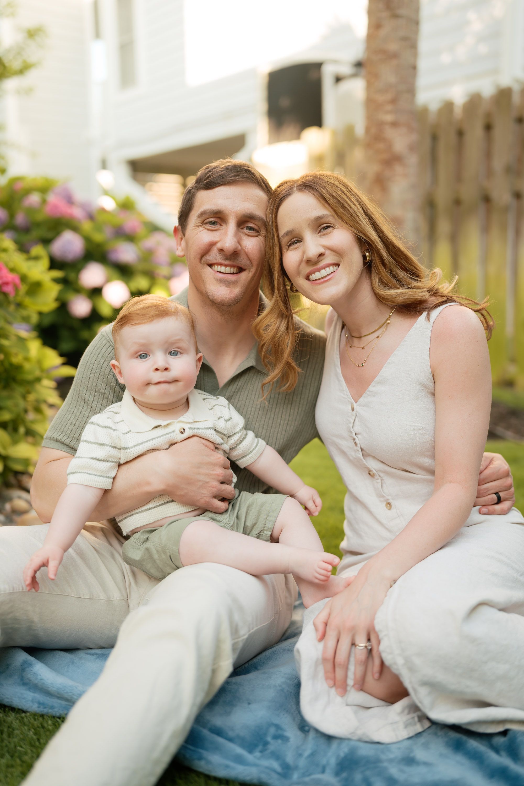Smiling couple with redhead baby sitting on lawn in garden setting.