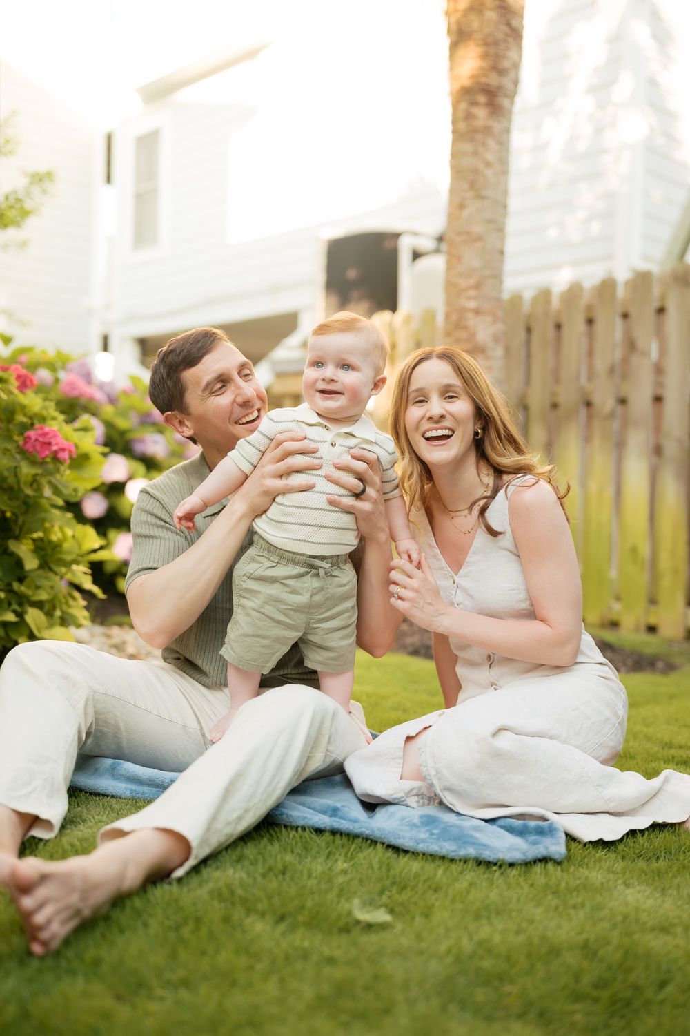 Happy young family with baby boy sitting on grass in backyard garden on sunny day.