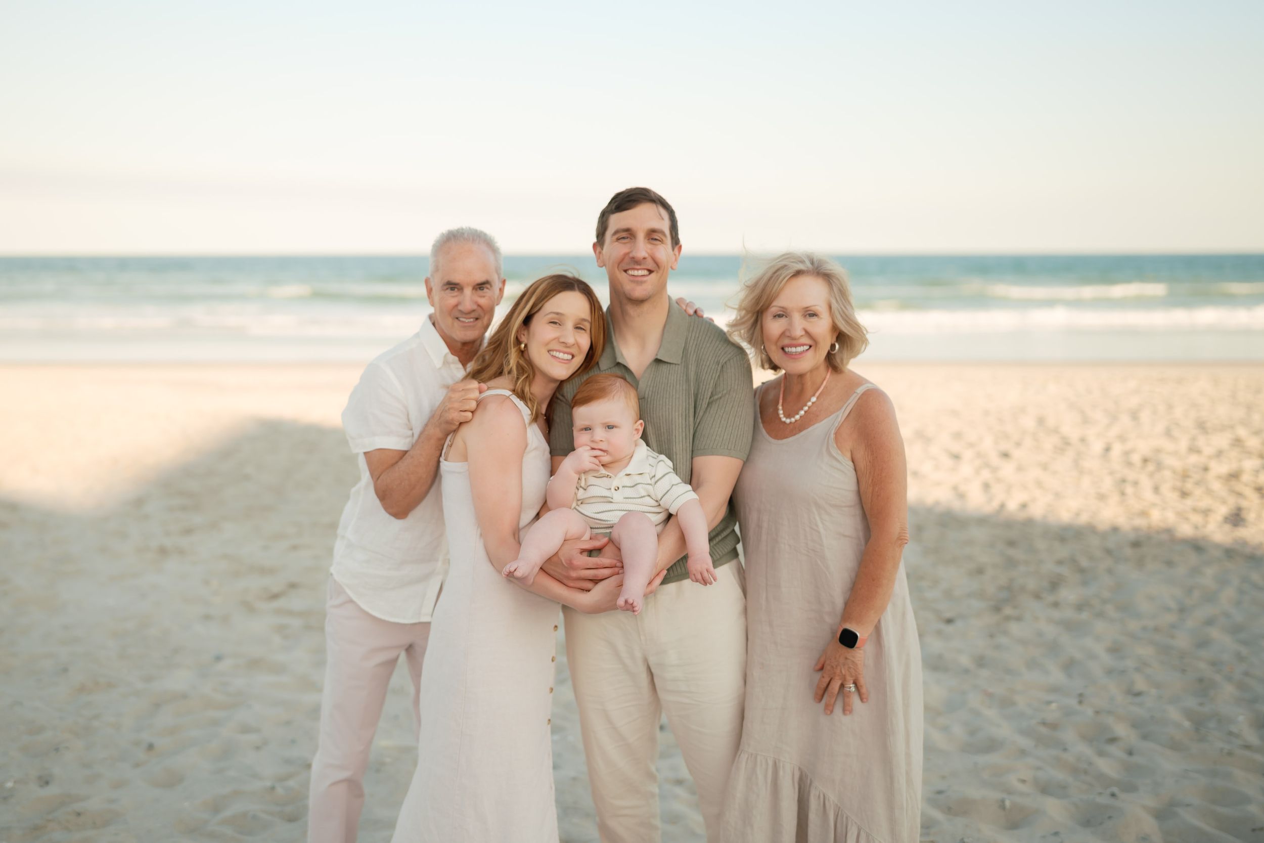 Family of five poses on a sandy beach at sunset, with parents holding a baby in the center.