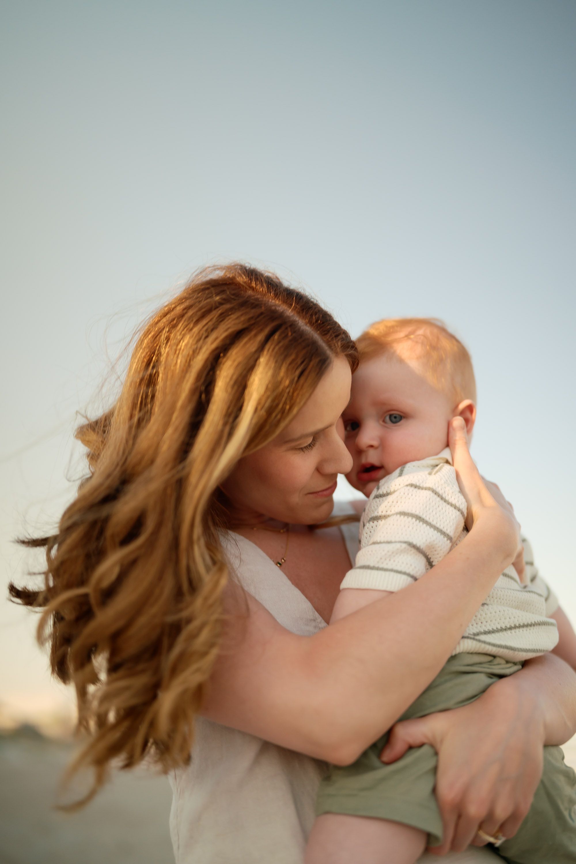 A mother with long red hair tenderly holds her baby outdoors at sunset.