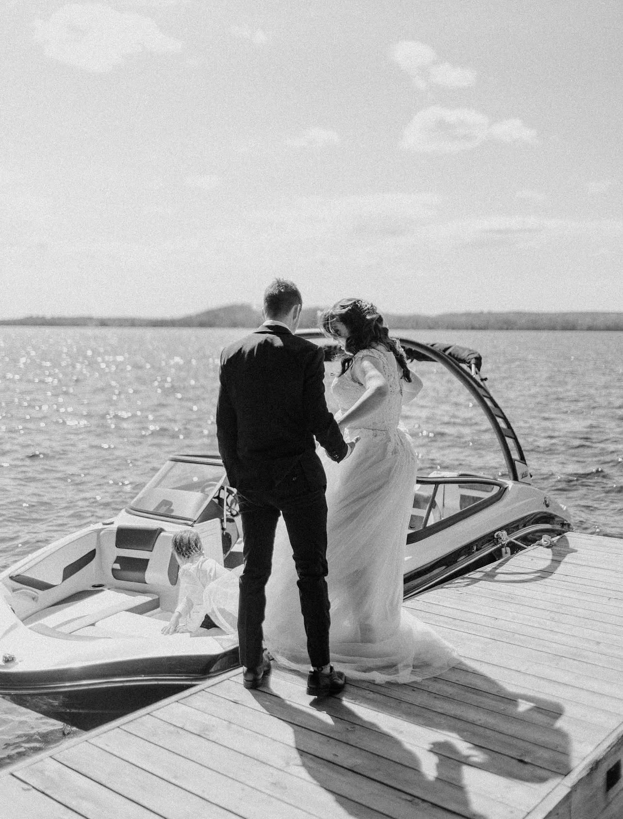 A couple stands on a wooden dock next to a sleek white speedboat on a tranquil lake with mountains in the distance.