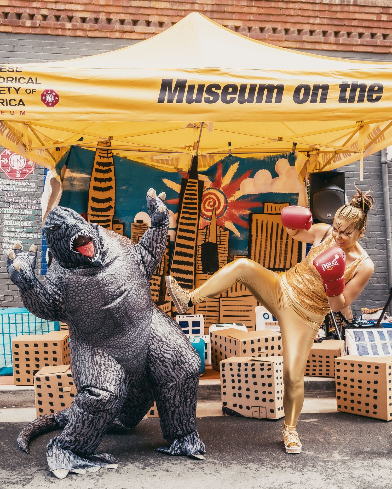 Woman in gold boxing outfit kicks near inflatable Godzilla costume at Museum on the Street outdoor event.