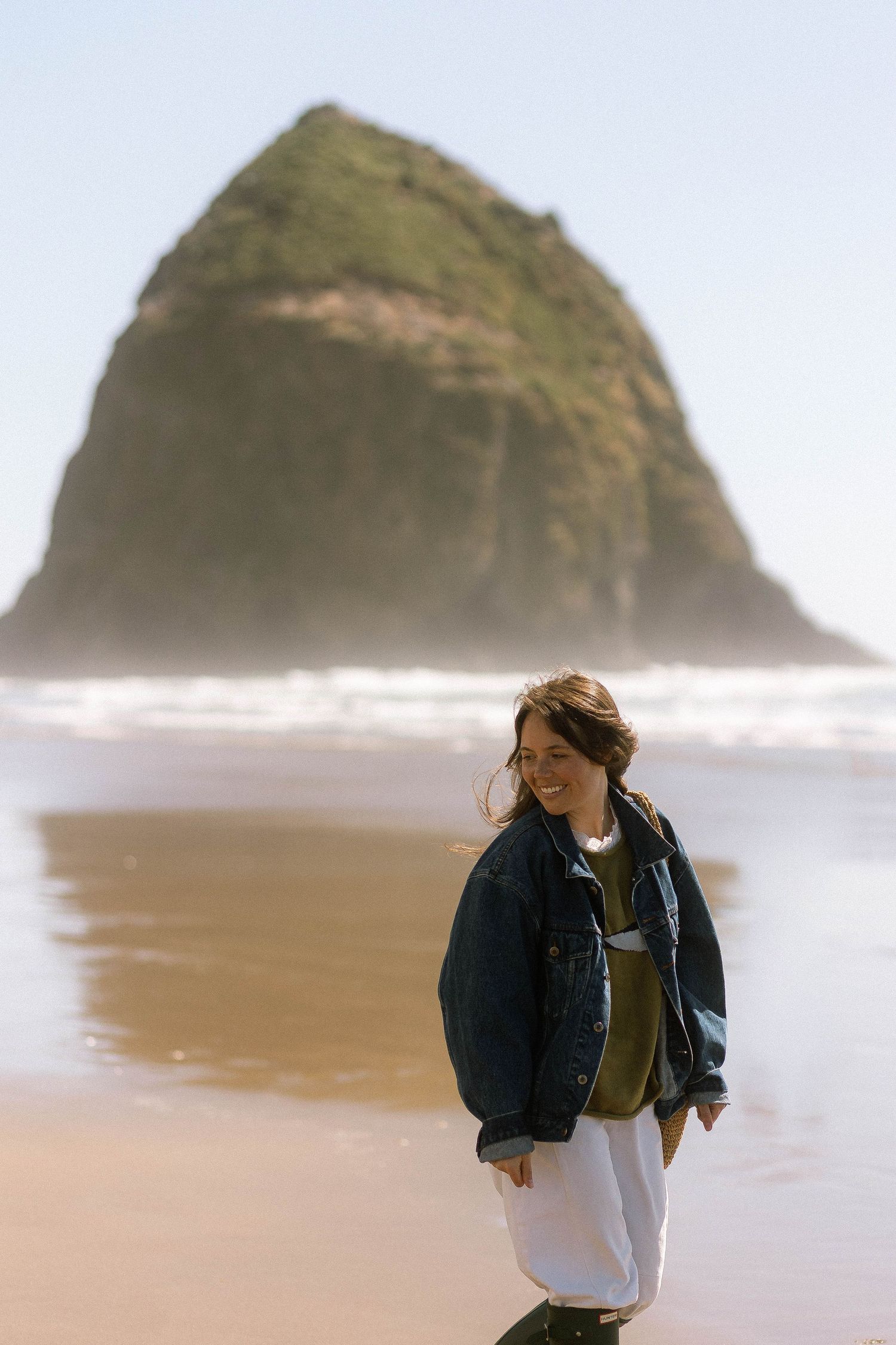 Smiling woman in denim jacket and white skirt standing on sandy beach with Haystack Rock in background.