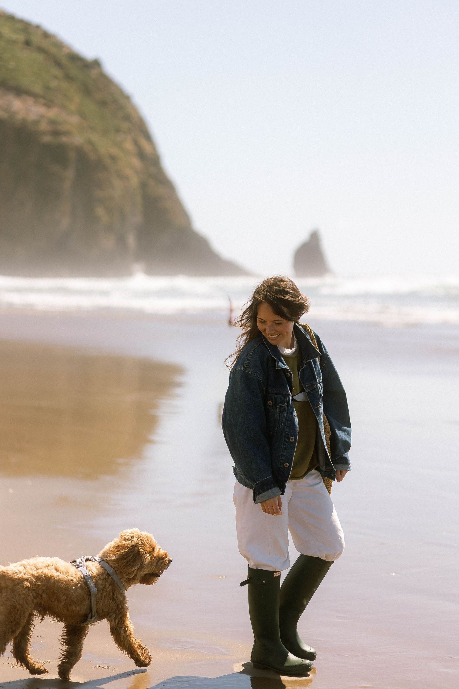 Woman in denim jacket and rain boots walks with a fluffy golden doodle on a misty beach with rocky cliffs.