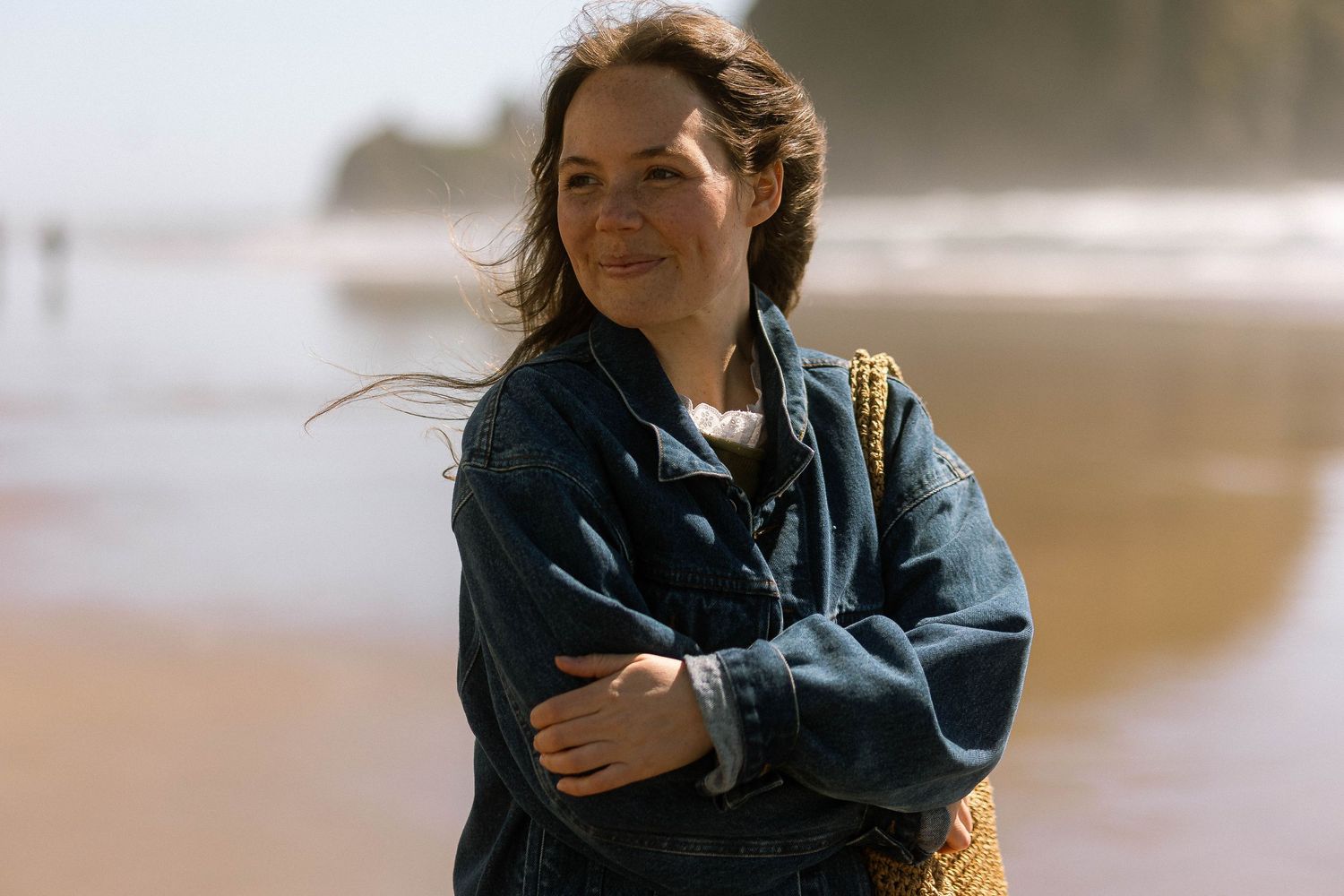 Woman in denim jacket standing on a sandy beach with arms crossed, smiling softly with ocean waves in the background.