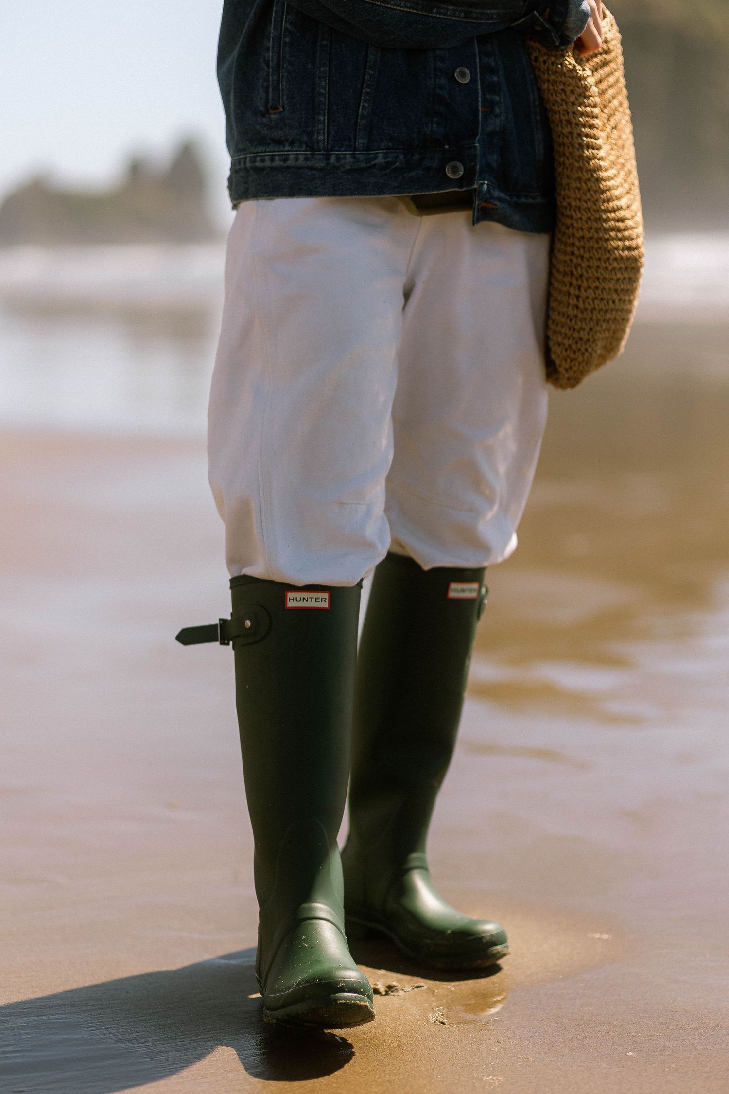 Woman wearing green Hunter rain boots, white pants, and denim jacket standing on wet sand at beach.