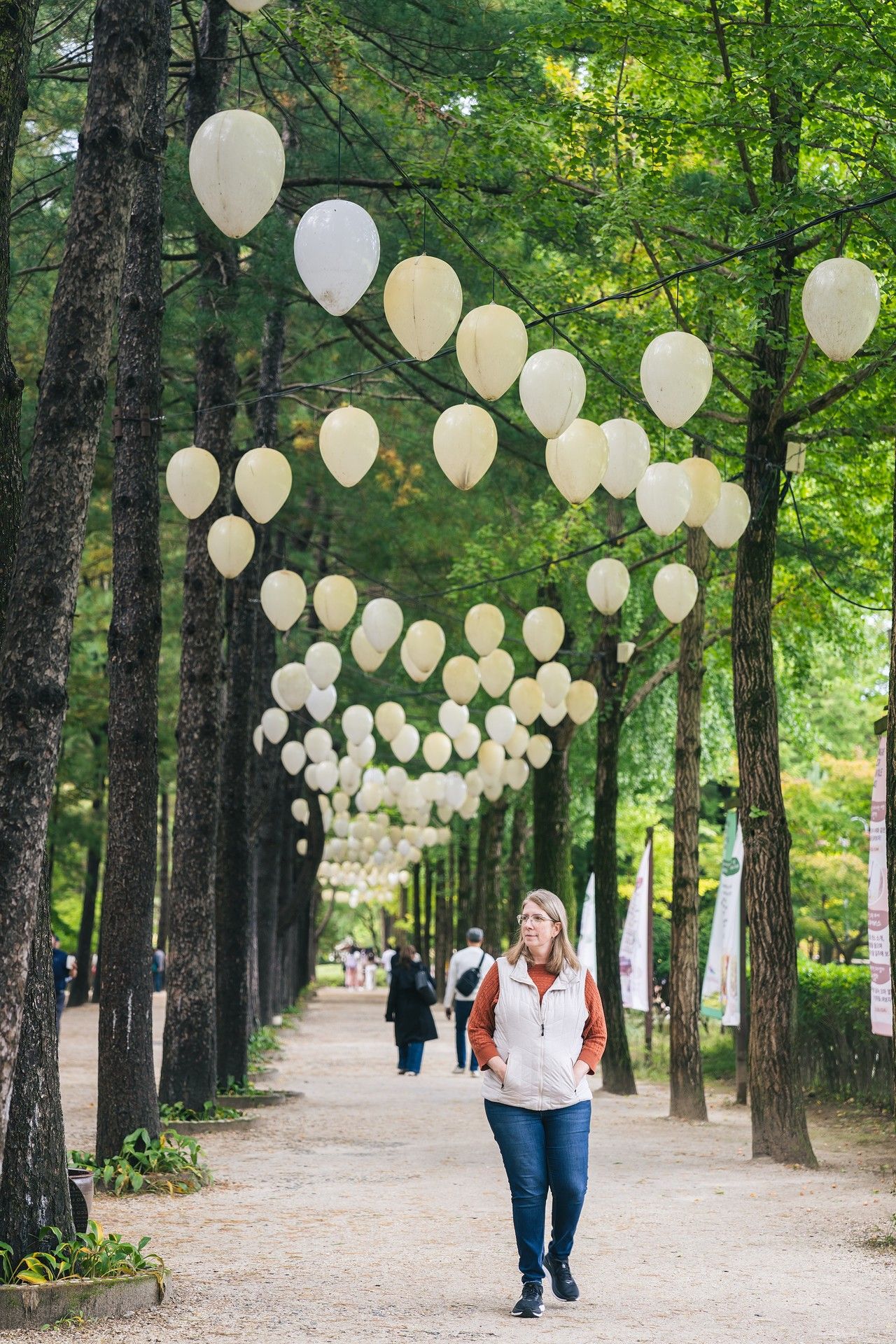 Woman walking along a tree-lined path decorated with white balloons in a serene park setting.