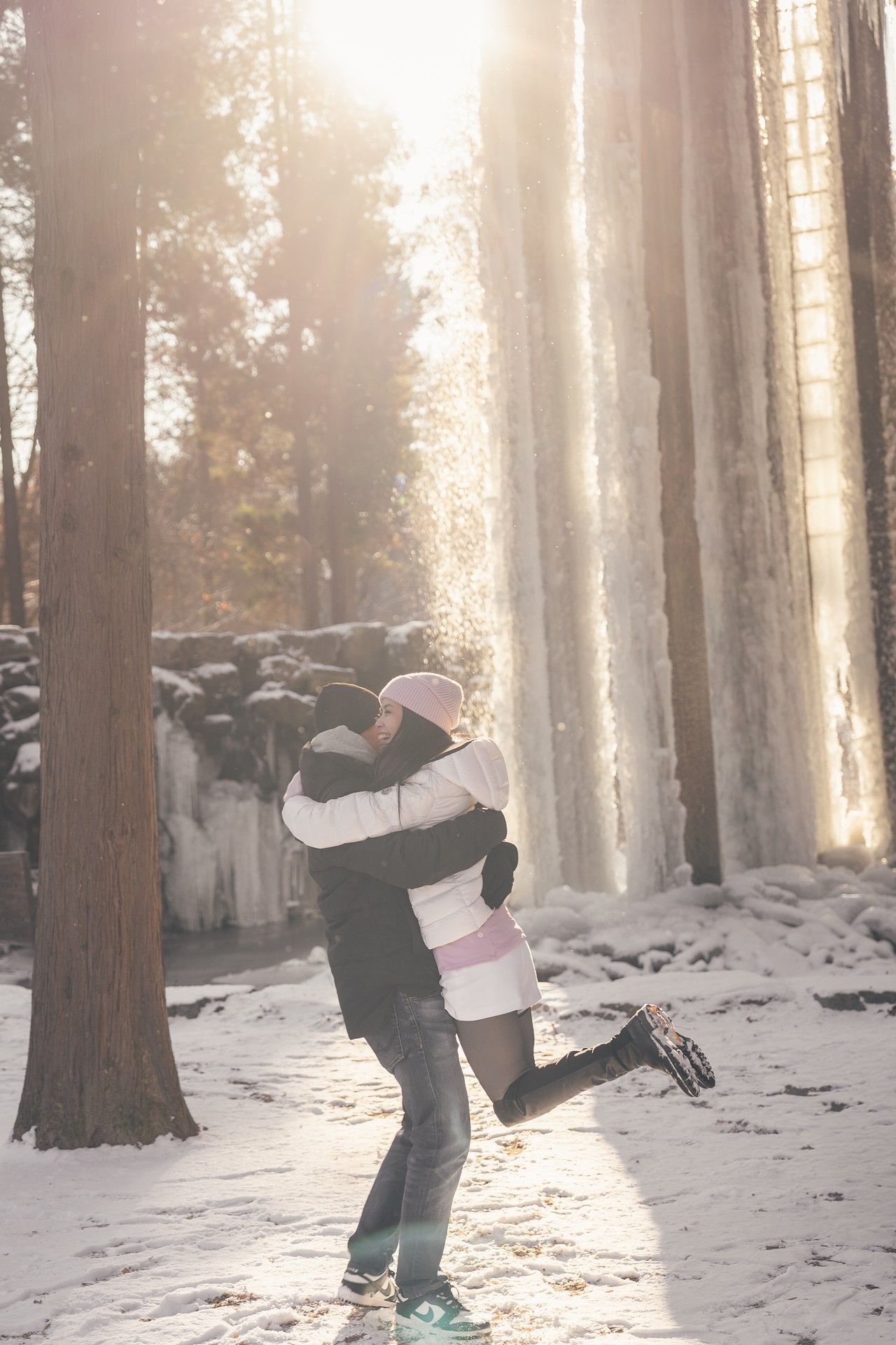 Couple embracing and lifting each other in a snowy winter forest with sunlight streaming through tall trees.