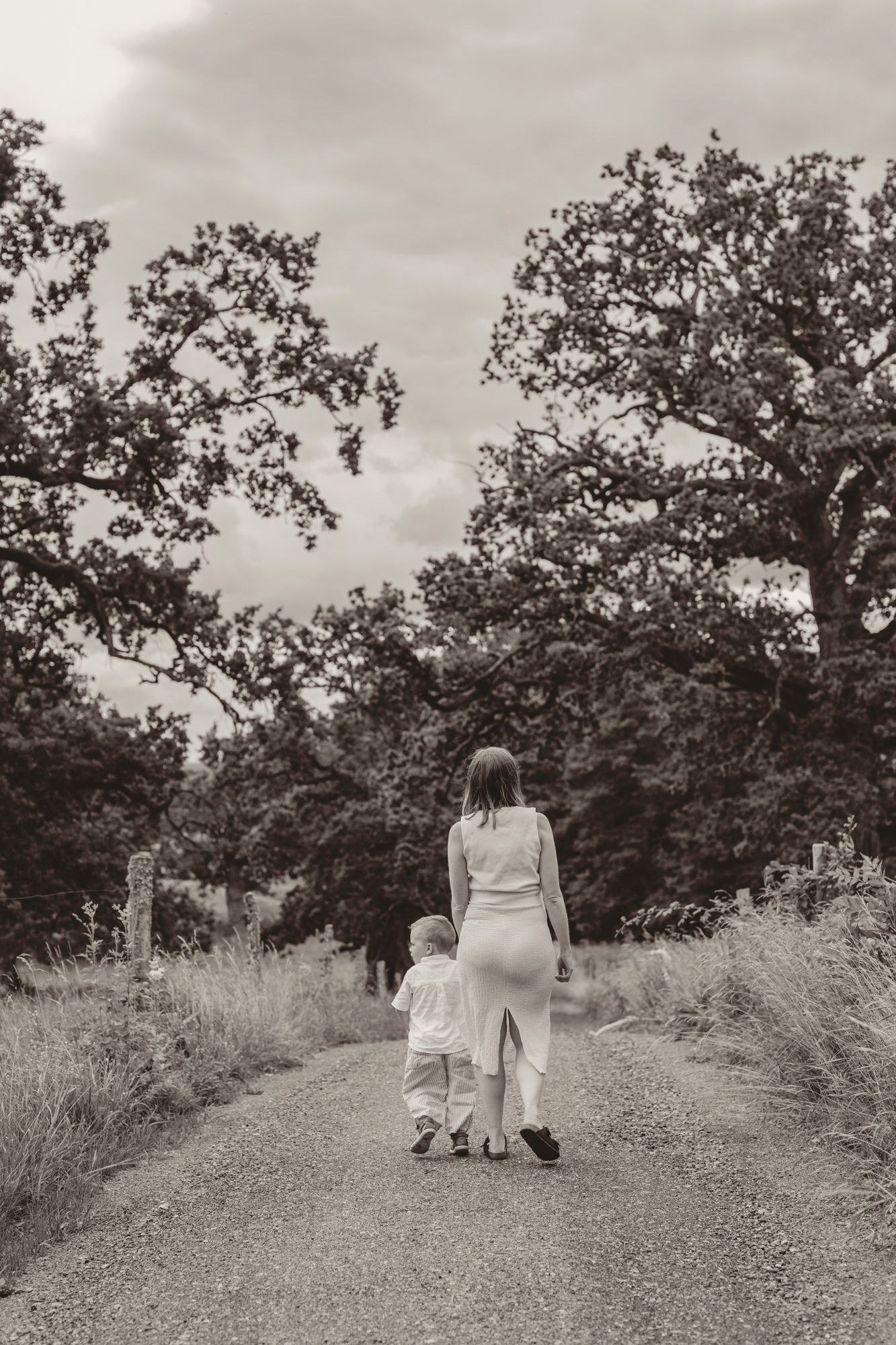 Black and white photo of a mother and toddler walking hand in hand down a rural path surrounded by trees.