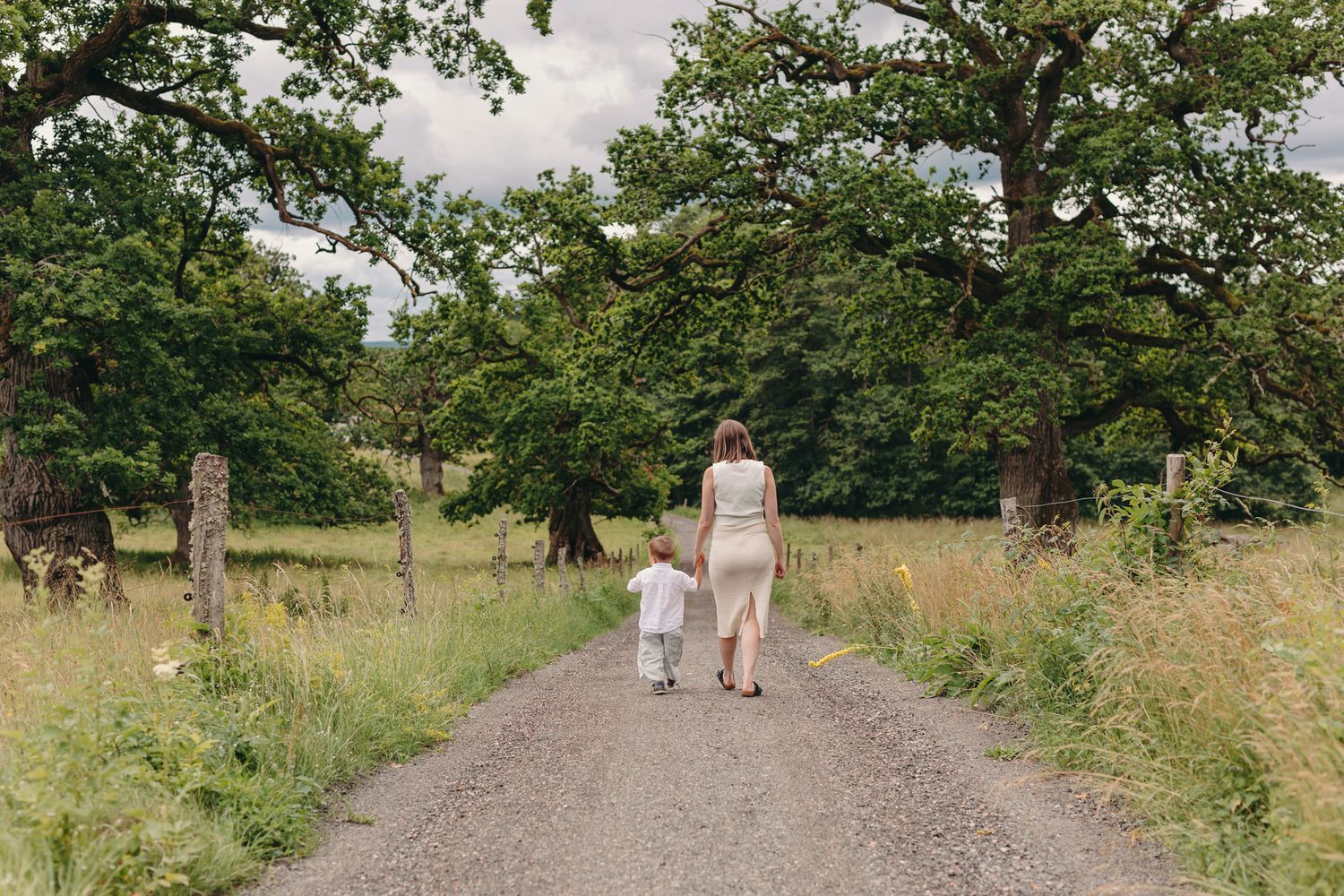 A mother and young child walk hand-in-hand down a rural path surrounded by lush green trees and meadows.