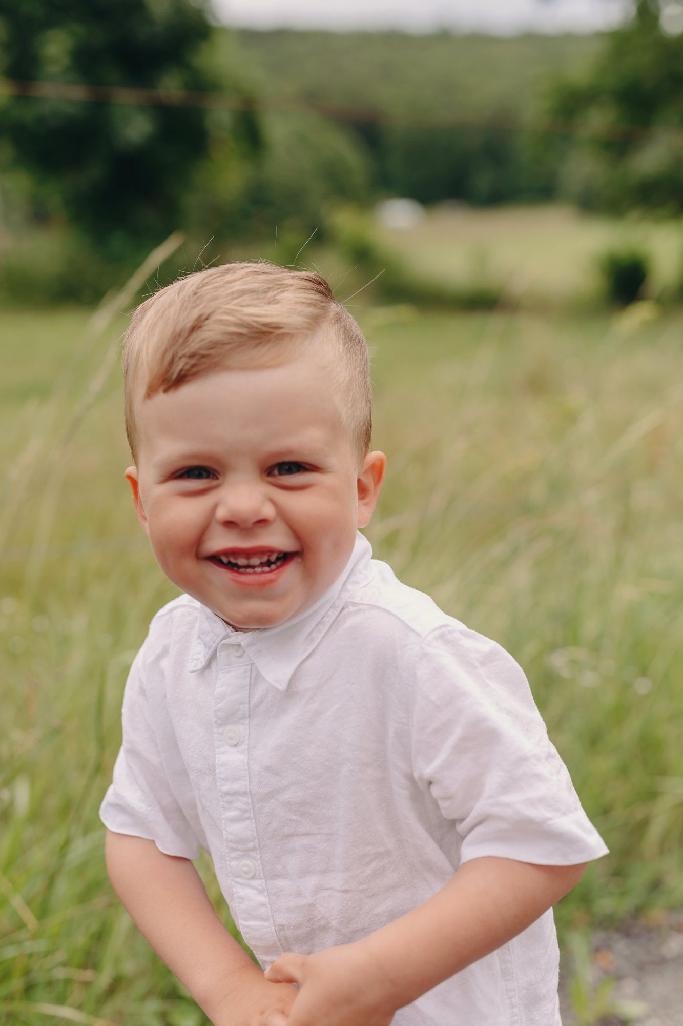 Happy smiling toddler boy in a white shirt laughing in a green grassy field outdoors.