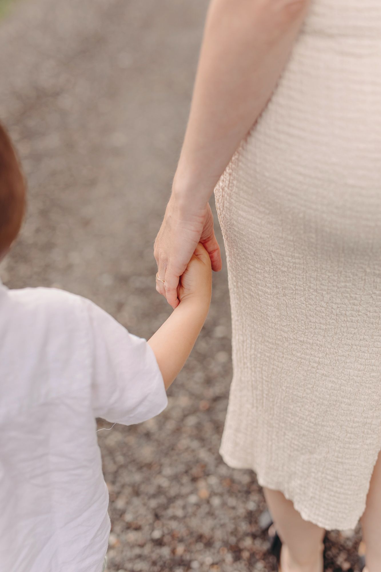 A mother and young child holding hands while walking on a gravel path, both wearing white outfits.