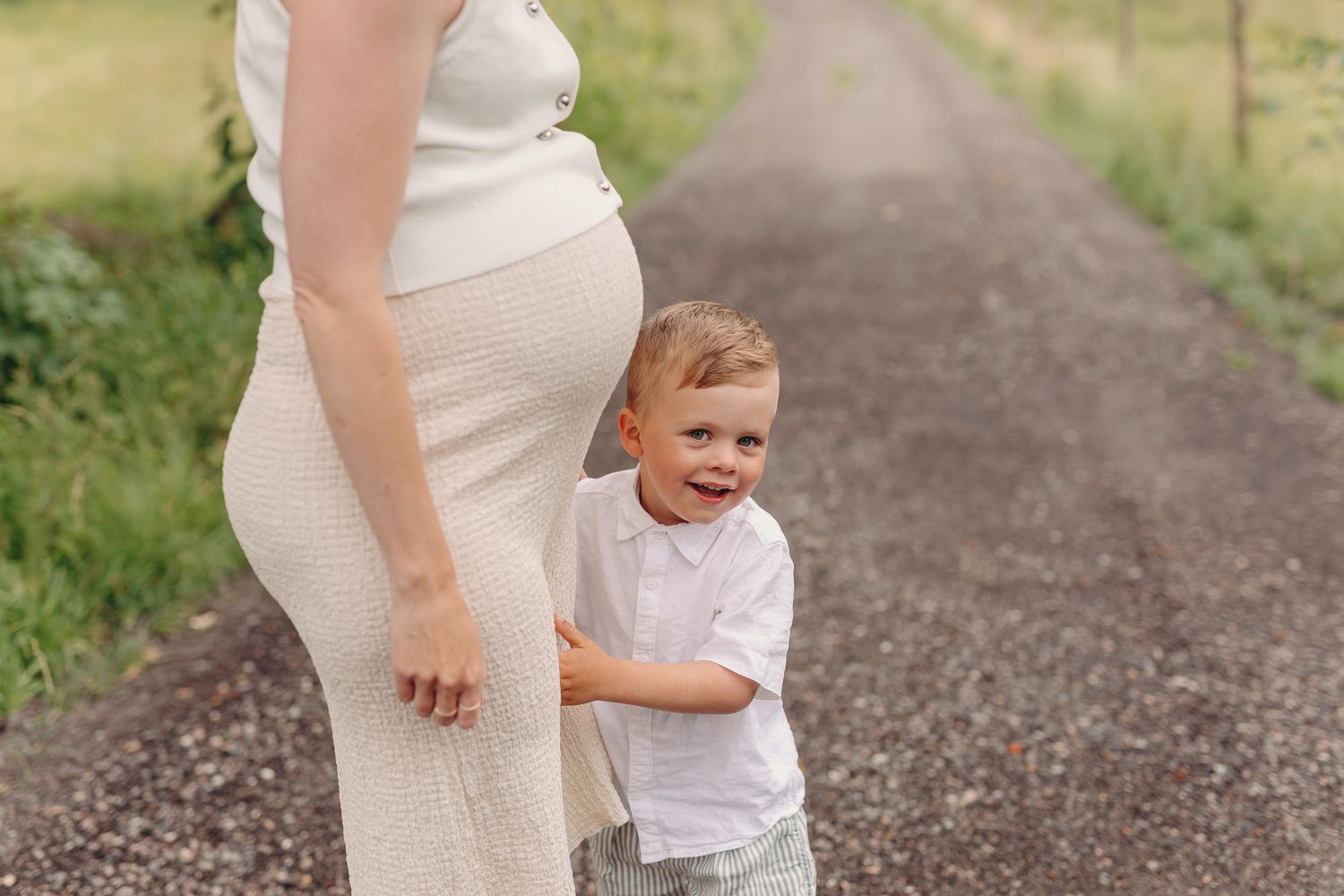 Toddler boy smiling beside pregnant mother on a scenic road, wearing white outfits in a lush green setting.
