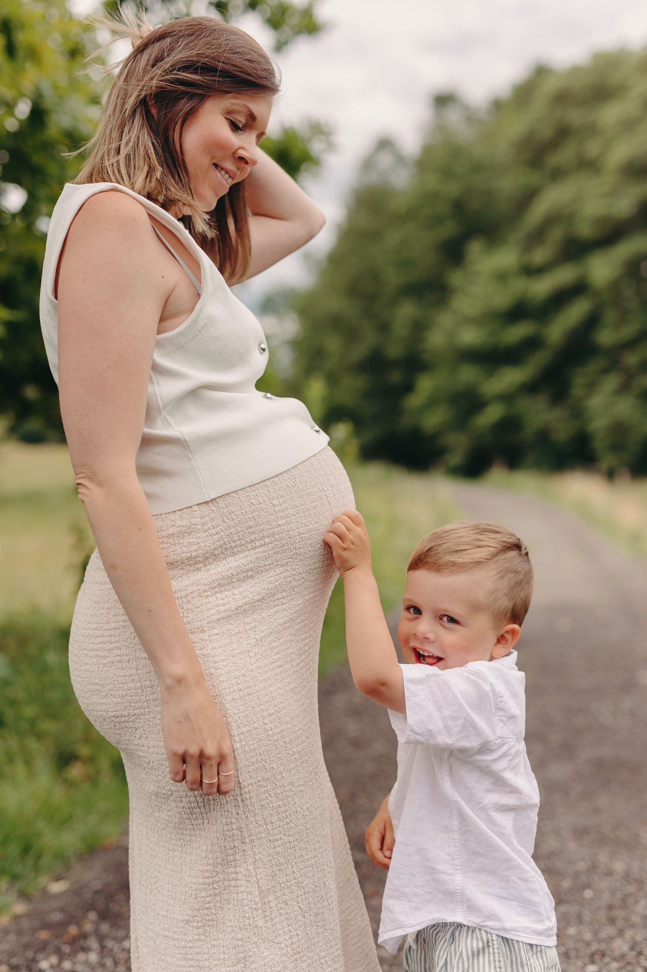 Pregnant woman in white dress smiling at toddler son touching her baby bump on a scenic path.