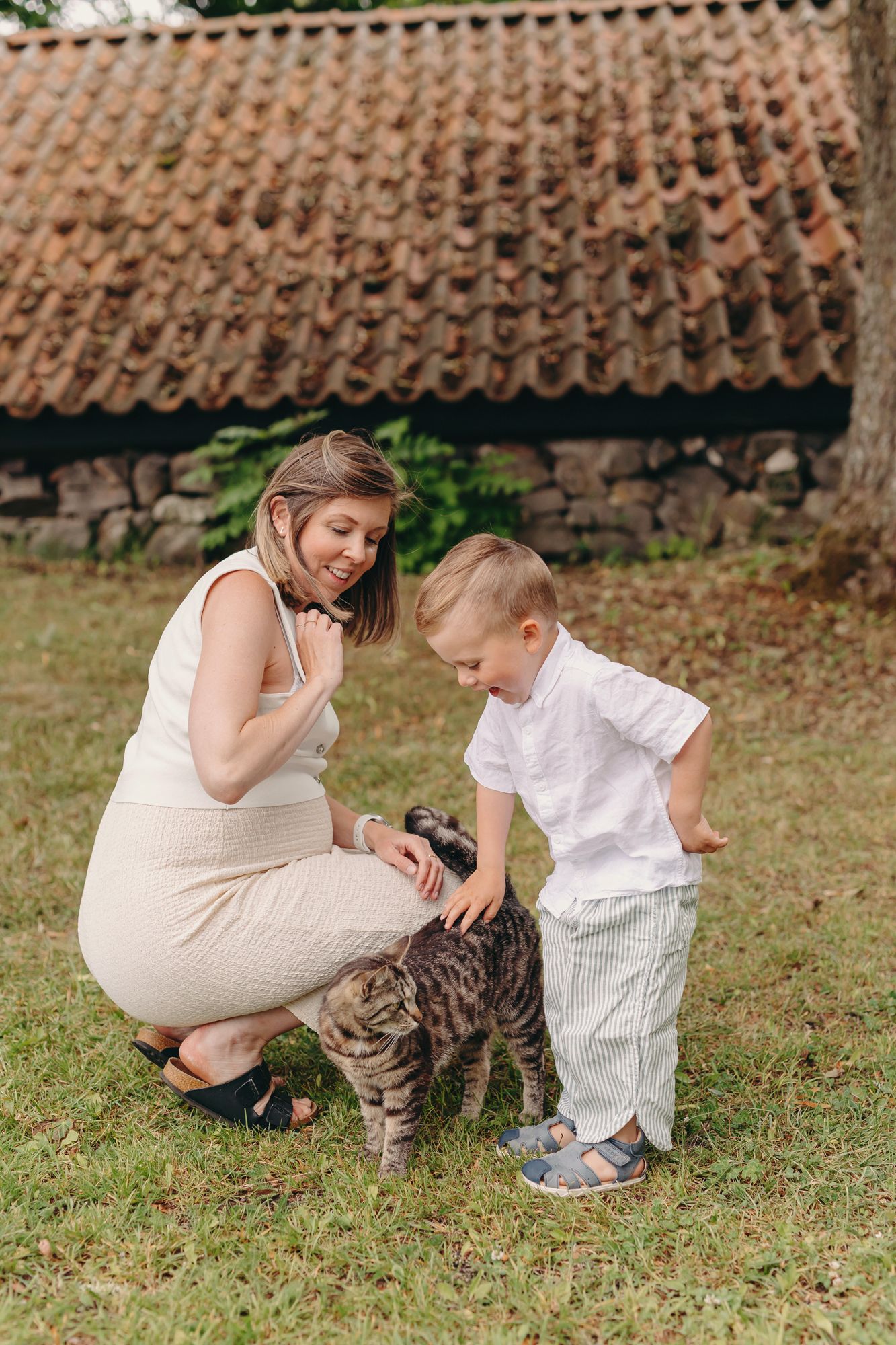 A smiling pregnant woman and young blonde boy gently pet a tabby cat outdoors near a rustic stone building.