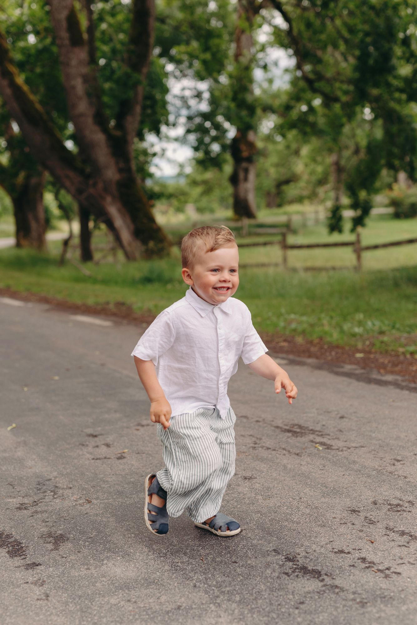A cheerful young boy in a white shirt and striped pants runs along a park path surrounded by green trees.