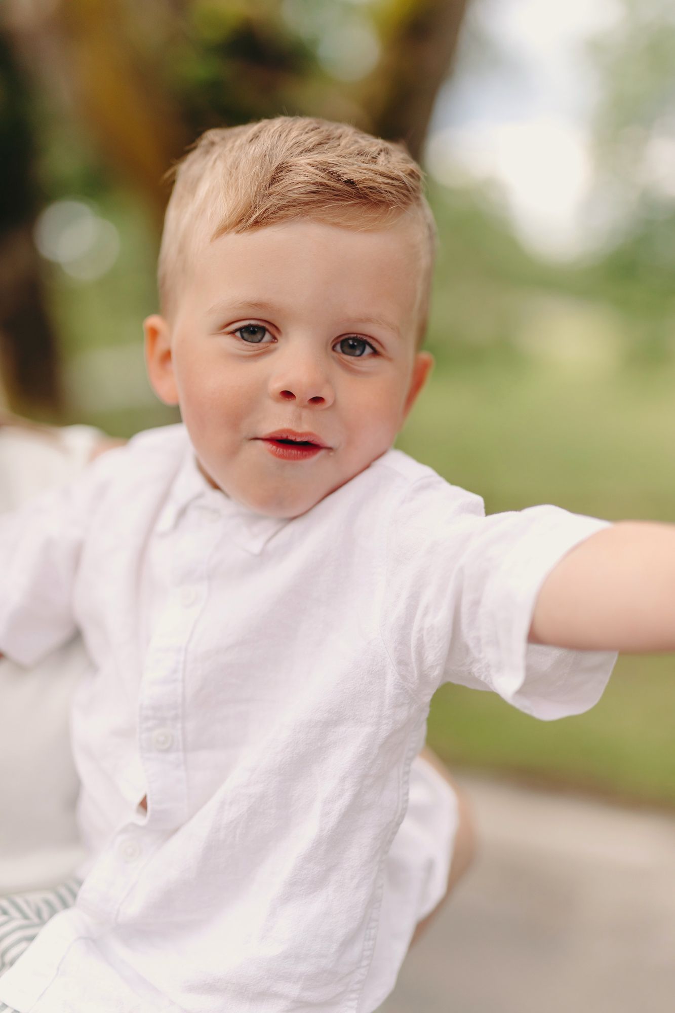 Cute toddler boy with blonde hair wearing a white linen shirt outdoors in a green park setting.
