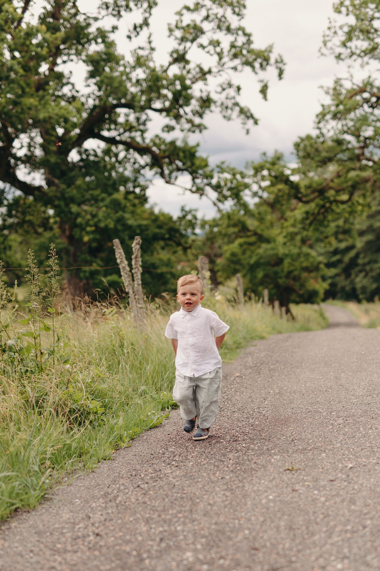 A young toddler boy in a white shirt walks along a rural road surrounded by lush green trees and grass.