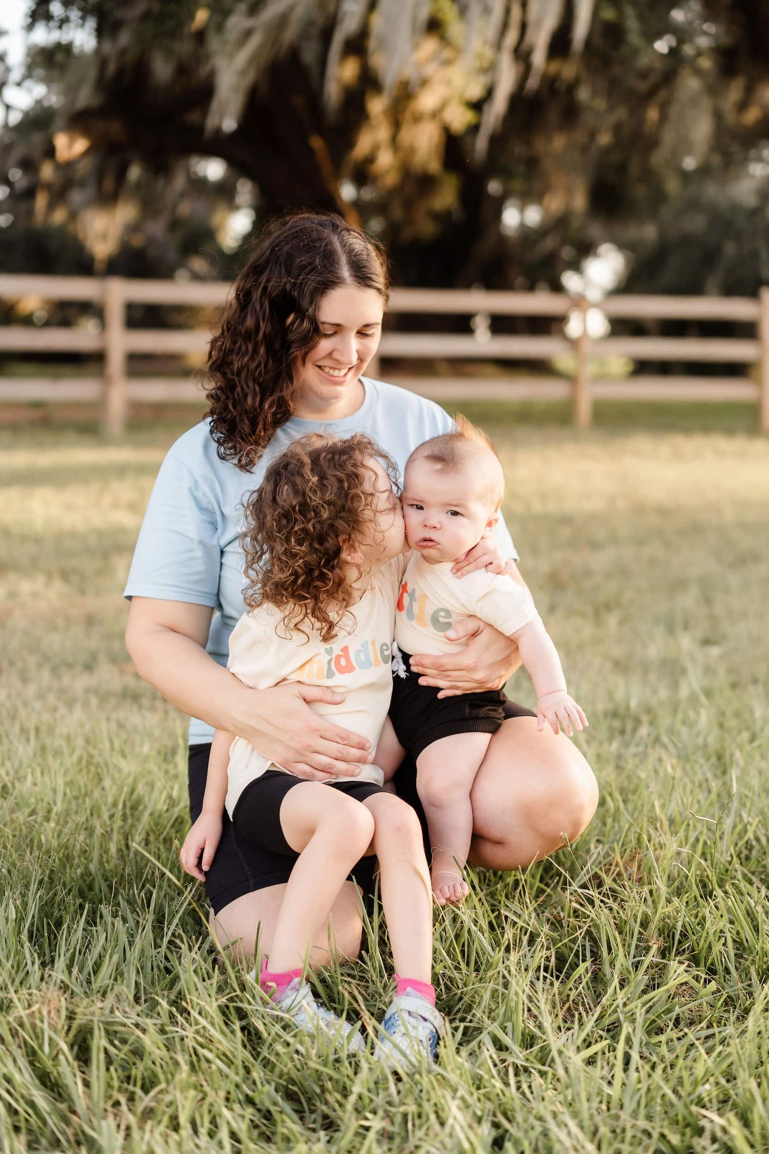 Parent in a red shirt lifts a giggling child wearing a red outfit up in the air at sunset in a grassy field.