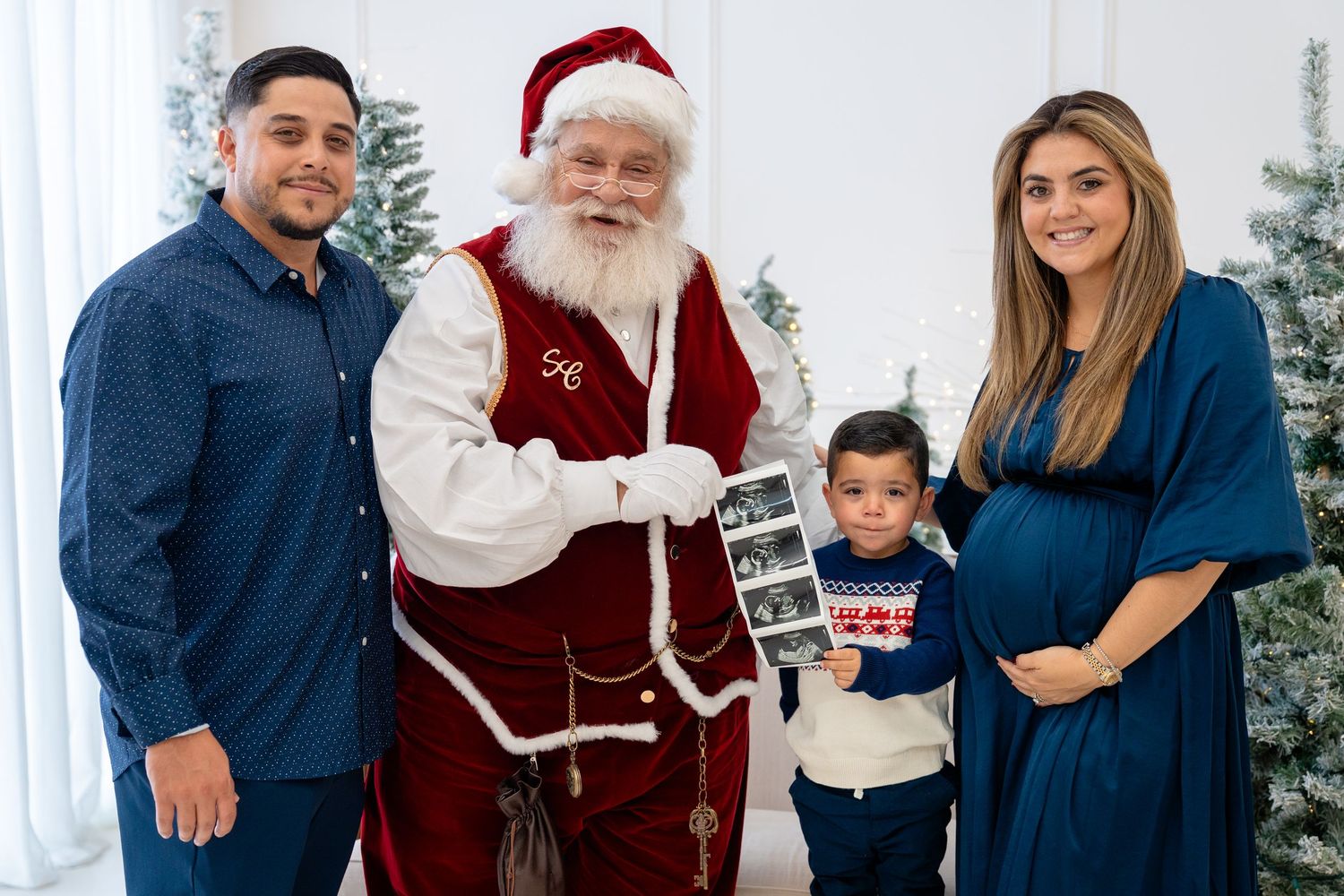 Family with Santa Claus holding ultrasound photos, pregnant woman, man, and toddler posing by Christmas trees.