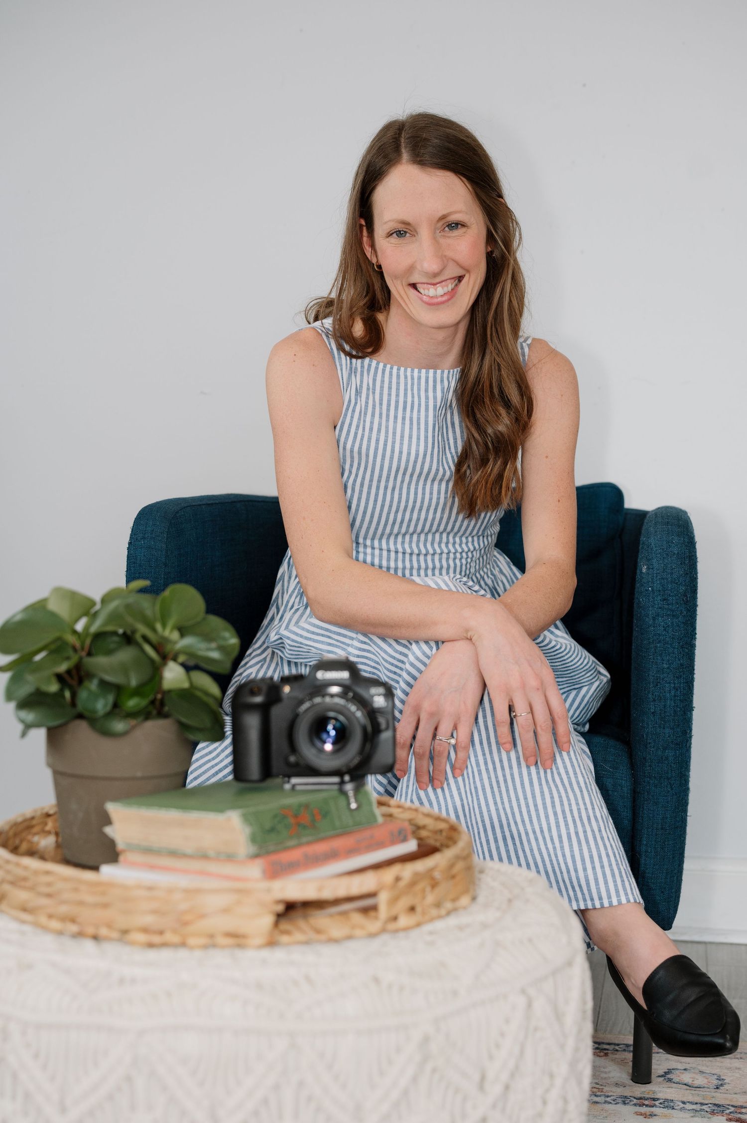 Woman in striped dress smiling in blue chair beside camera, books, and plant on wicker tray.