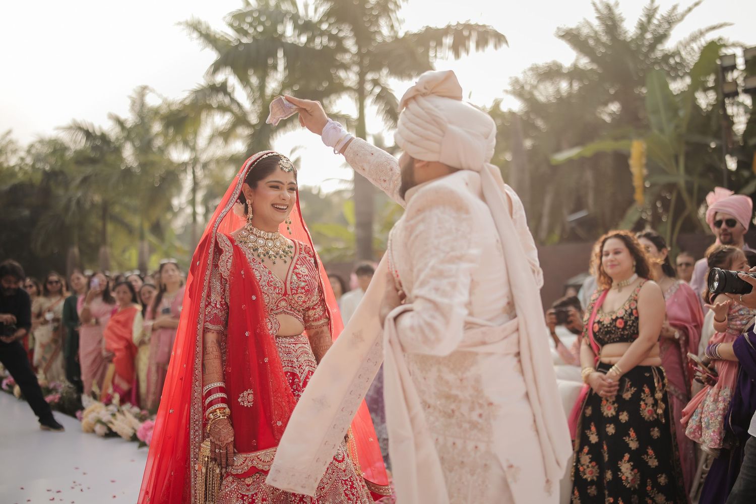 Indian bride in red lehenga and groom in white sherwani dancing at outdoor wedding ceremony with palm trees.