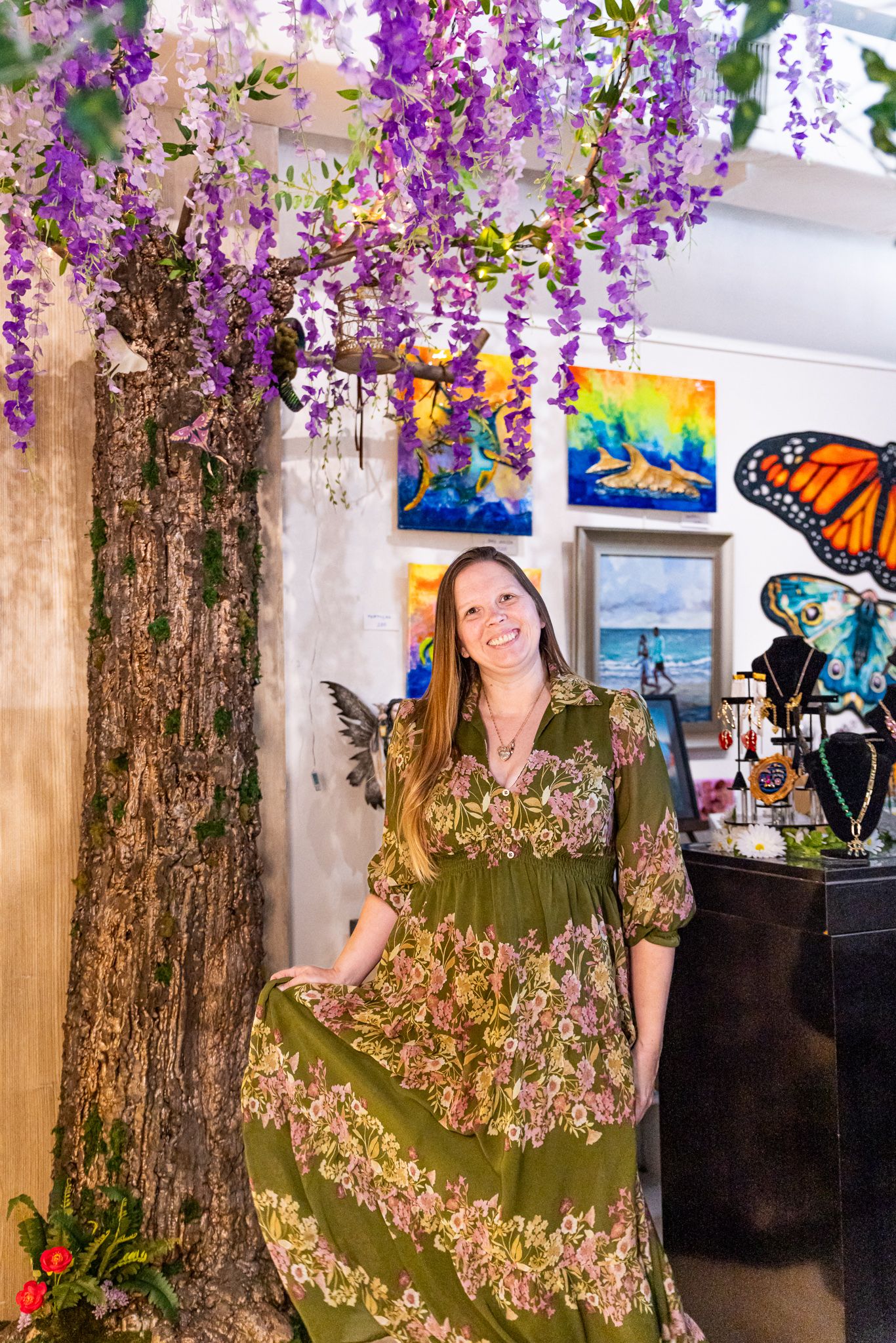 Woman in floral green dress smiling under purple wisteria tree in colorful art gallery with butterfly paintings.