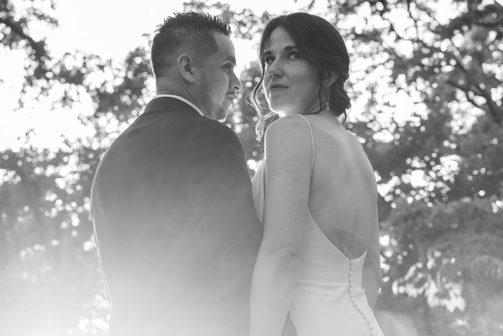 Black and white wedding photo of bride and groom standing together outdoors under trees, bride glancing back over shoulder.