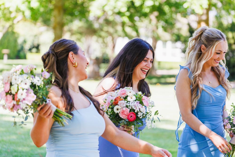 Three bridesmaids in blue dresses laughing and holding pink floral bouquets outdoors at a wedding.
