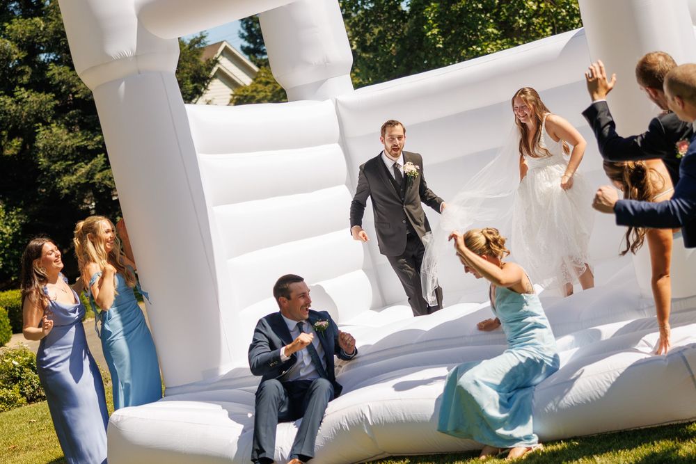 Bride and groom joyfully bouncing in a white inflatable bounce house with wedding guests at an outdoor reception.