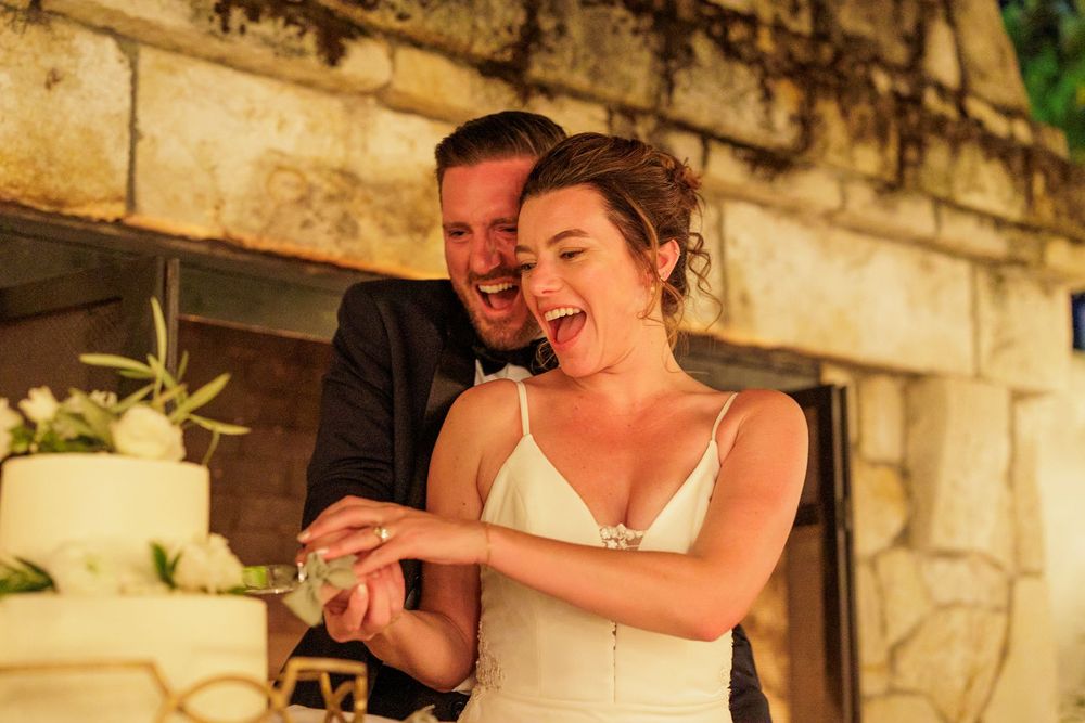 Bride and groom laughing joyfully while cutting their white wedding cake at an outdoor stone venue.