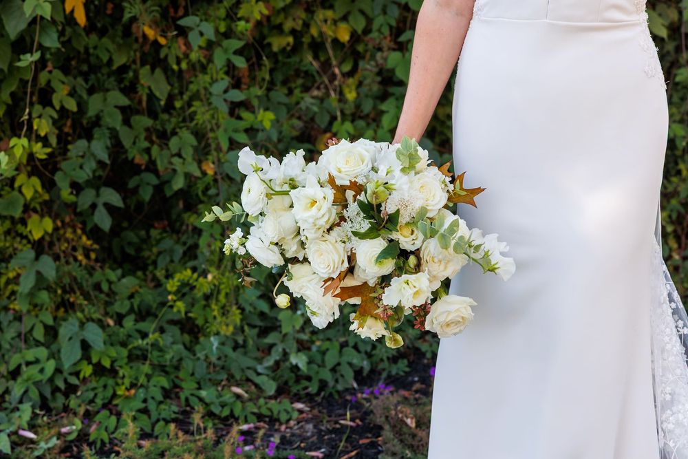 Bride in white gown holding lush white floral bouquet with greenery outdoors against ivy-covered wall.