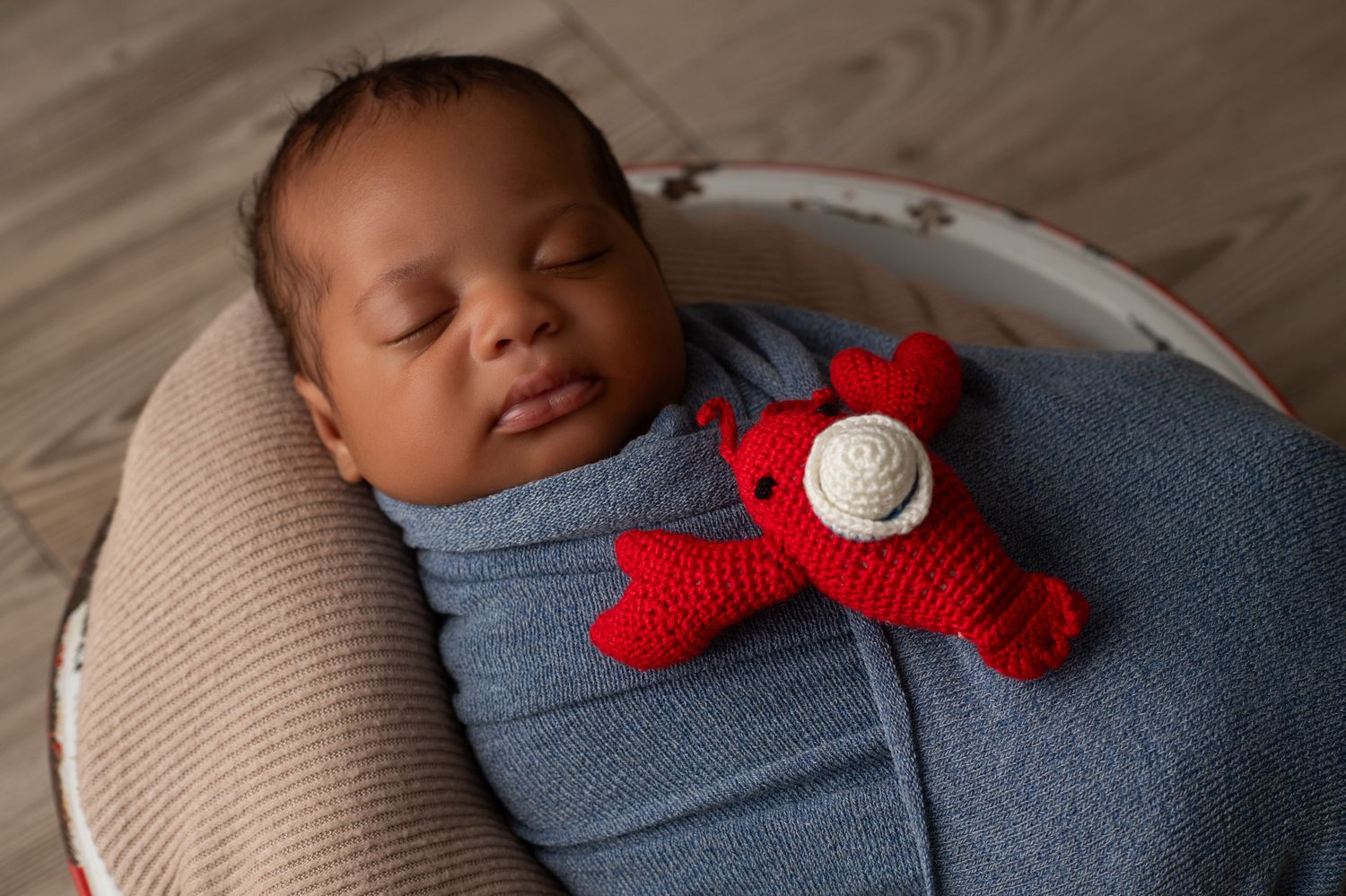 Sleeping newborn baby swaddled in blue wrap, resting in a bowl with a red crocheted lobster toy.