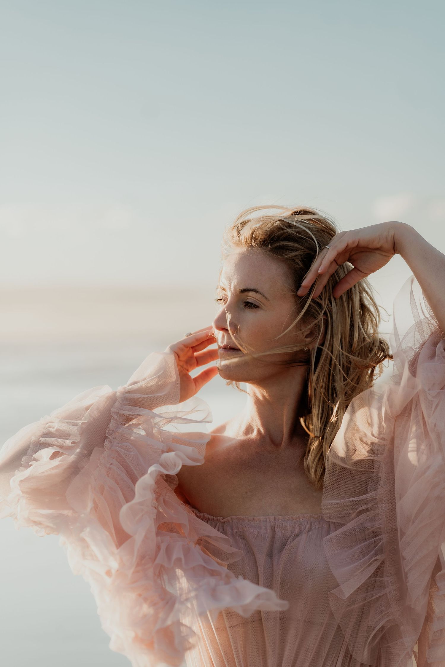 Woman in flowing pink tulle dress poses gracefully outdoors, hair blowing in wind against soft sky backdrop.