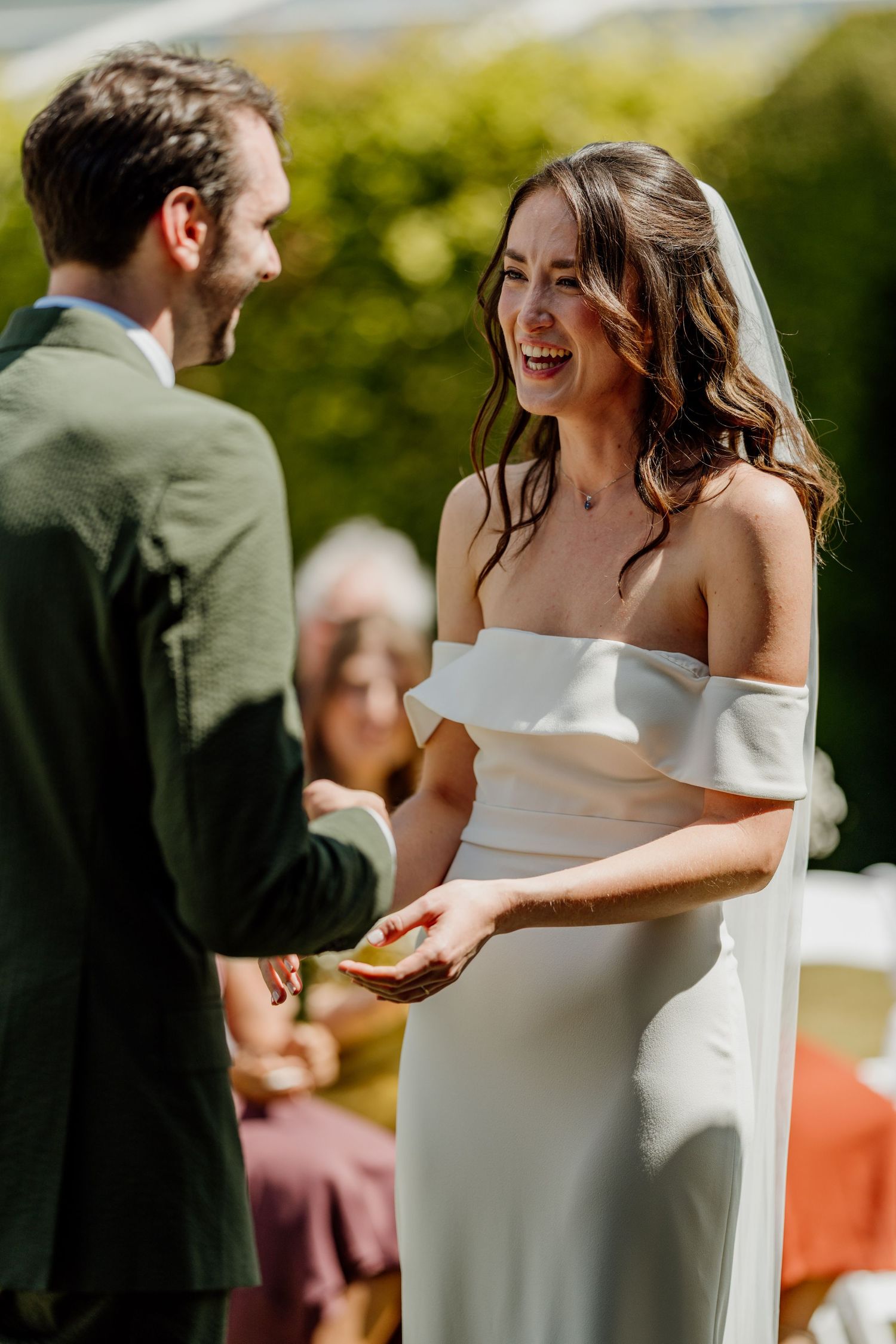 Bride in off-shoulder white gown laughs joyfully during outdoor wedding ceremony while holding rings at Kingston Estate in Devon.
