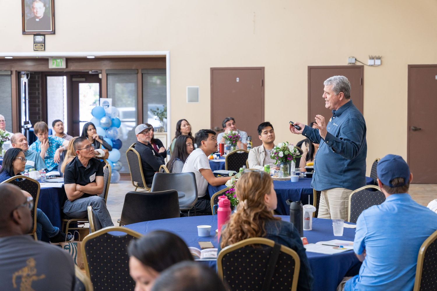 Speaker addresses diverse group of attendees seated at round tables during a community event with blue balloon decorations.