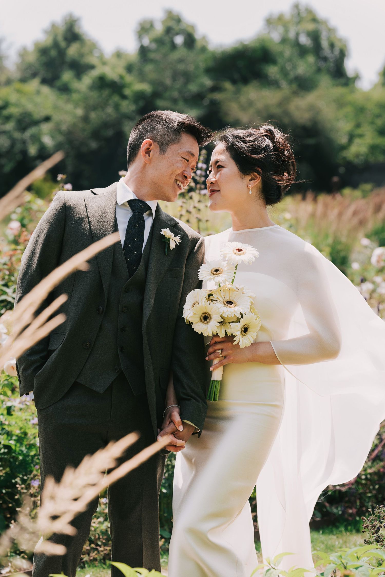 A smiling bride and groom share a romantic moment at Greenwich Park London she holds white gerbera daisy bouquet, wearing elegant cape gown.