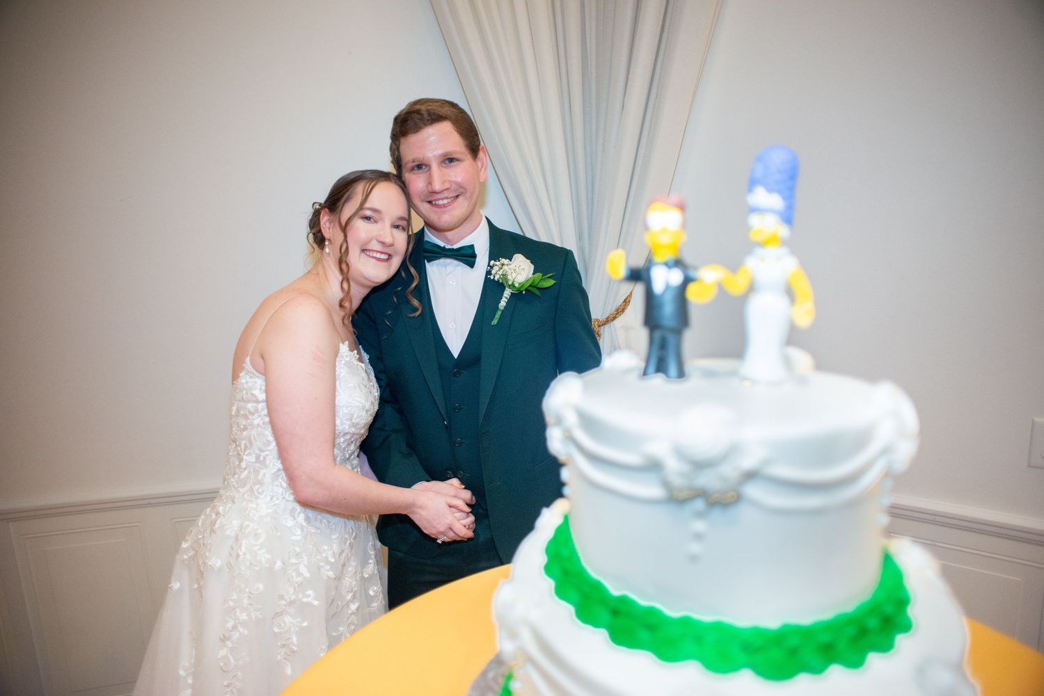 Newlyweds smile beside their wedding cake topped with Simpsons figurines as cake toppers.
