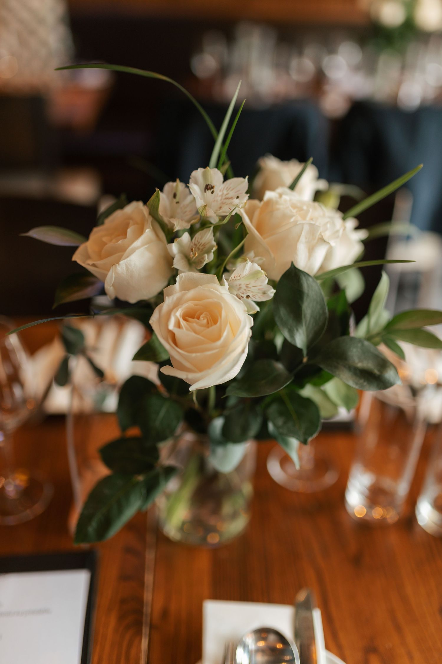 Elegant wedding centerpiece with cream roses and white flowers in a glass vase on a wooden reception table.