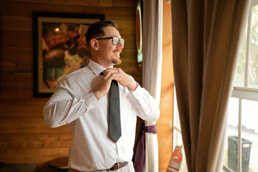 Groom in white dress shirt and glasses adjusting his gray tie near a window in a rustic wood-paneled room.