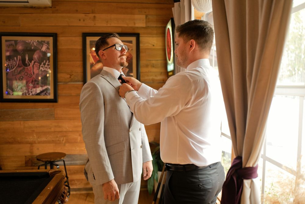 A groomsman helps adjust the groom's tie while both men wear white dress shirts in a rustic wood-paneled room.