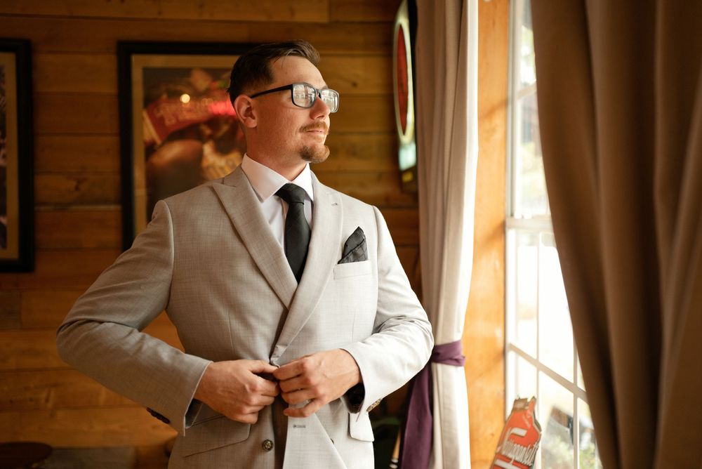 Groom in light gray suit adjusting jacket buttons by window with natural light in rustic wood-paneled venue.